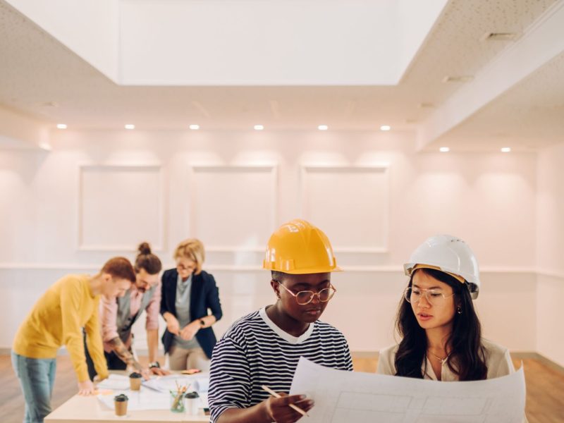 Portrait of two multiracial architect looking at blueprints in an office Two female architect looking at blueprint at architecture studio. Portrait of african american and vietnamese woman engineer in office with their colleagues having a meeting in the background.